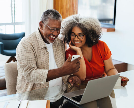 smiling mature couple at home on mobile phone and laptop