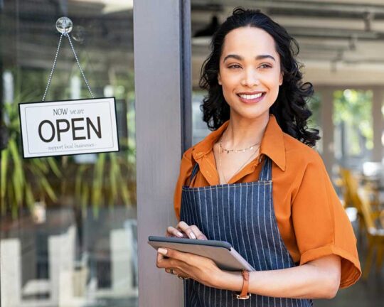 smiling female business owner standing outside door with open sign